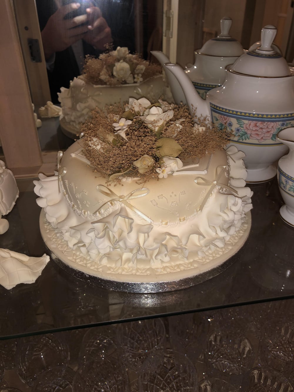 A white cake decorated with ruffled icing, small bows, and topped with dried flowers, sits on a reflective surface beside elegant china teapots and cups.