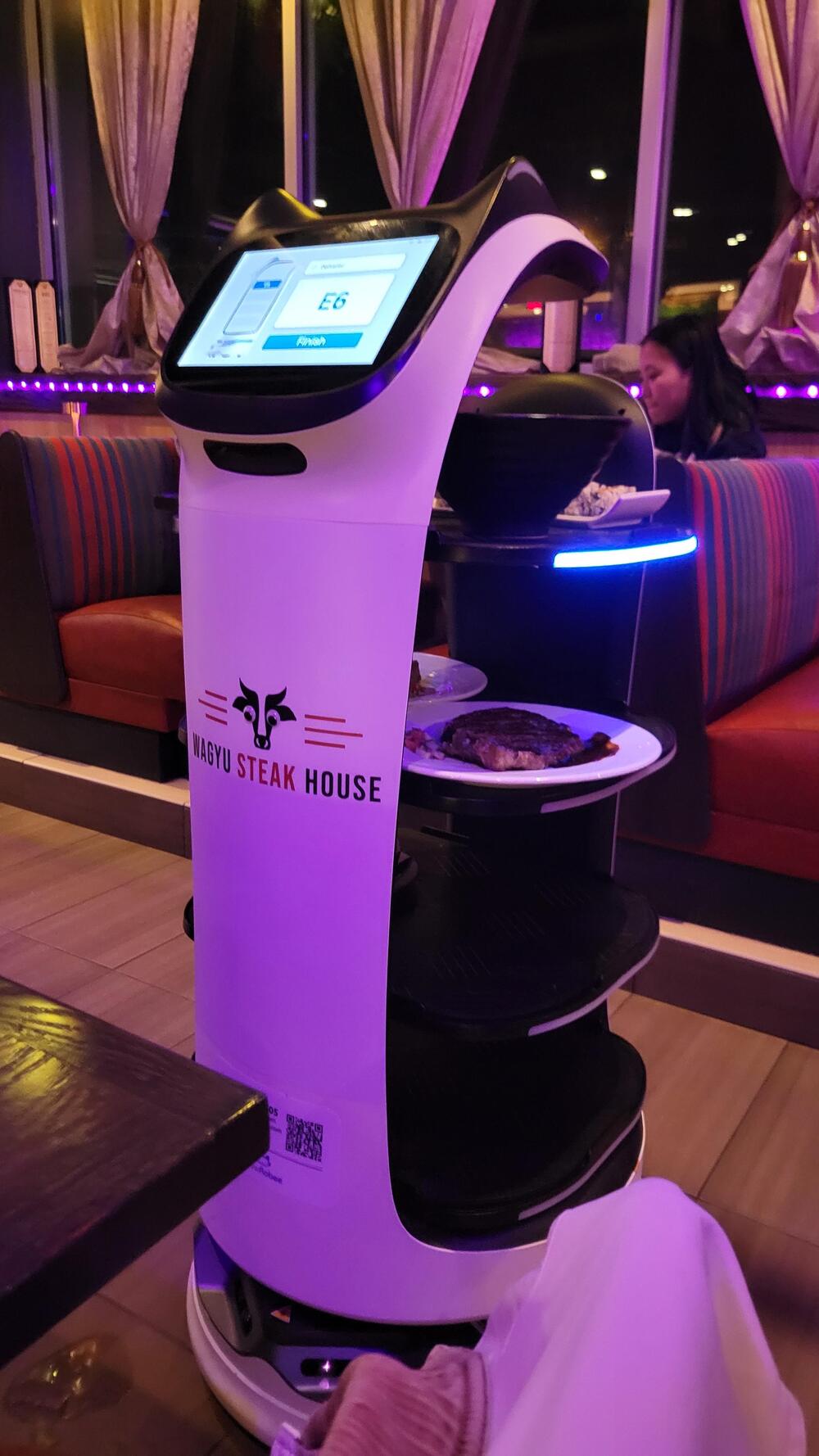 A robot server with shelves carrying food stands in a restaurant called Wagyu Steak House. Diners are seated in booths behind it.