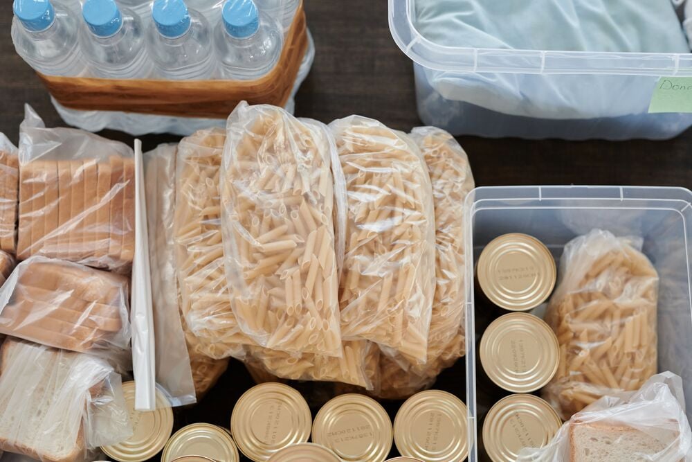 Clear containers and bags hold penne pasta, canned food, sliced bread, and bottles of water, arranged on a dark surface, suggesting food storage or distribution.