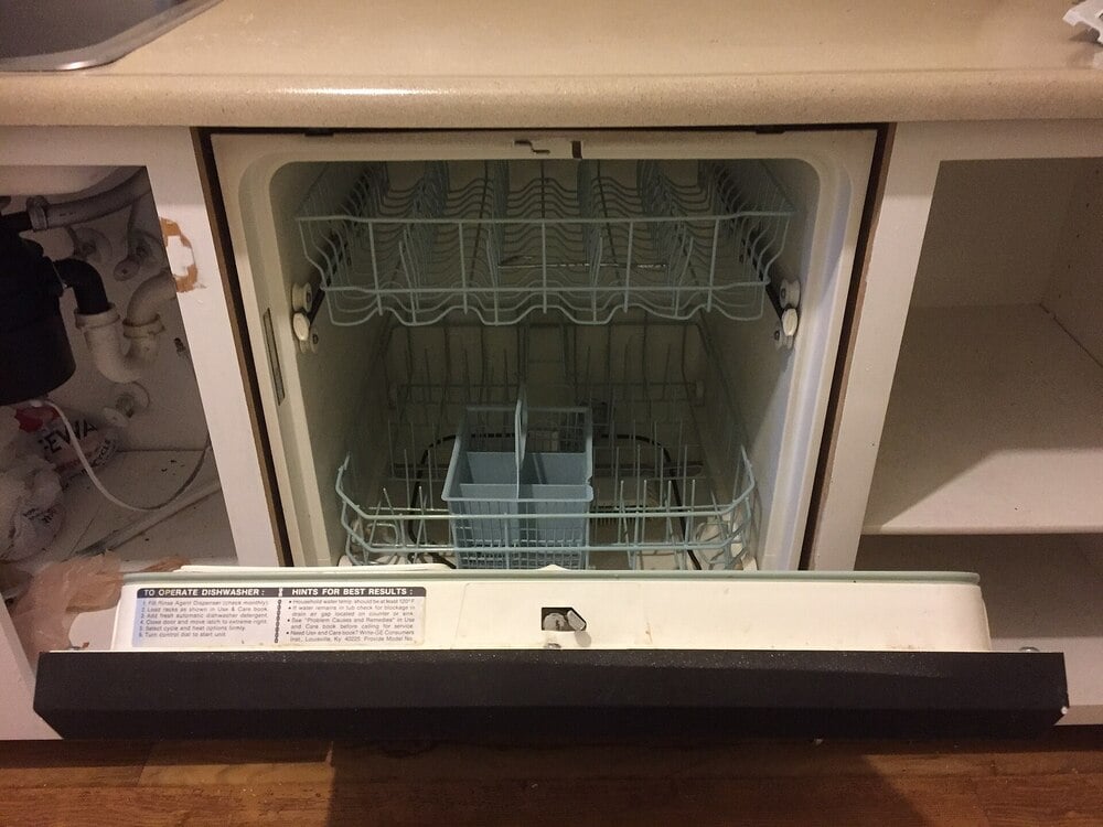 An open, empty dishwasher is built into a kitchen cabinet. The lower and upper racks are visible, along with the utensil holder. The dishwasher door is fully open, resting on a wooden floor.