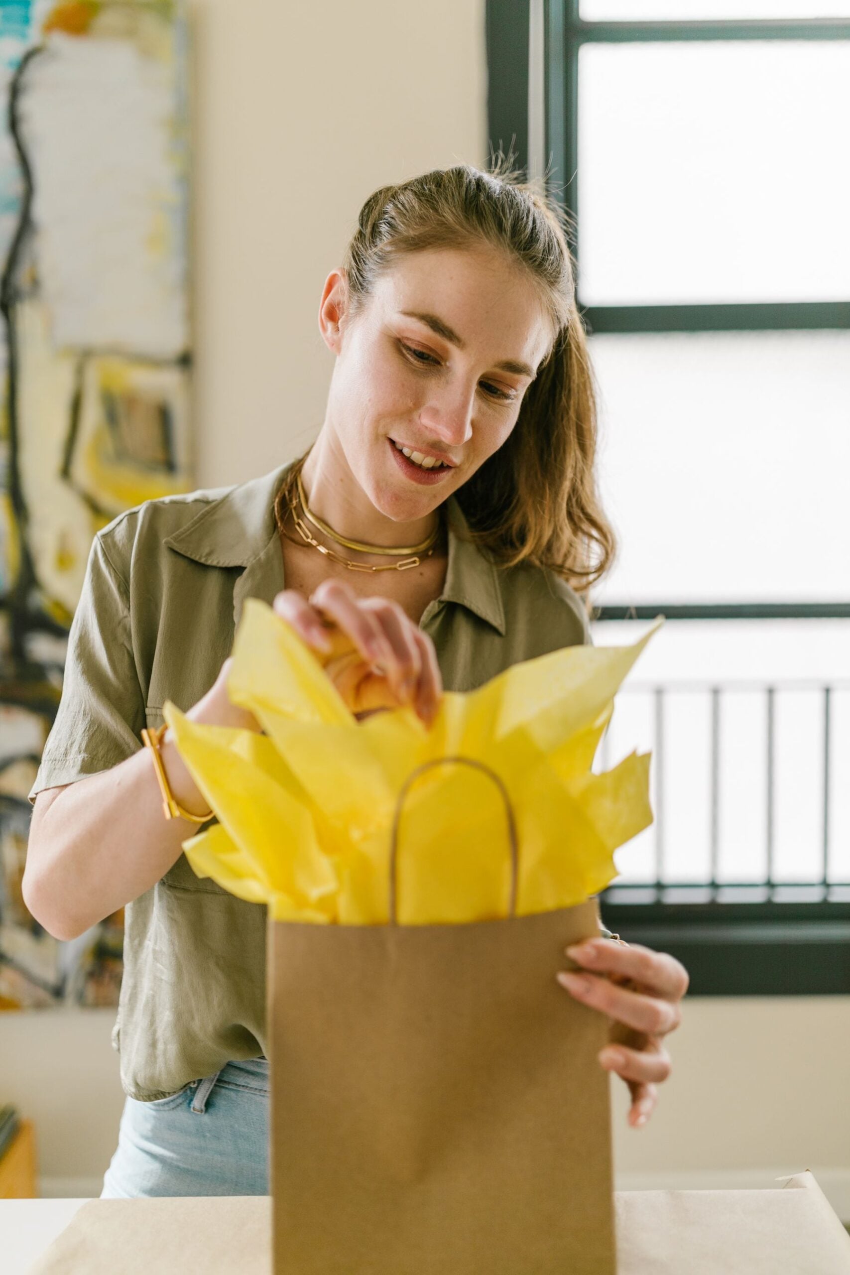 A person smiles while arranging yellow tissue paper in a brown gift bag, standing indoors near a window with natural light.