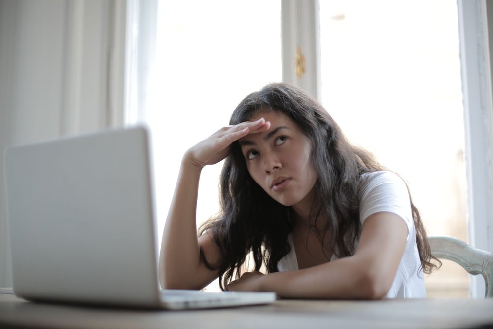 Woman with long hair sits at a desk with a laptop, looking concerned or frustrated, resting her head on one hand while the other shields her eyes, with a bright window in the background.