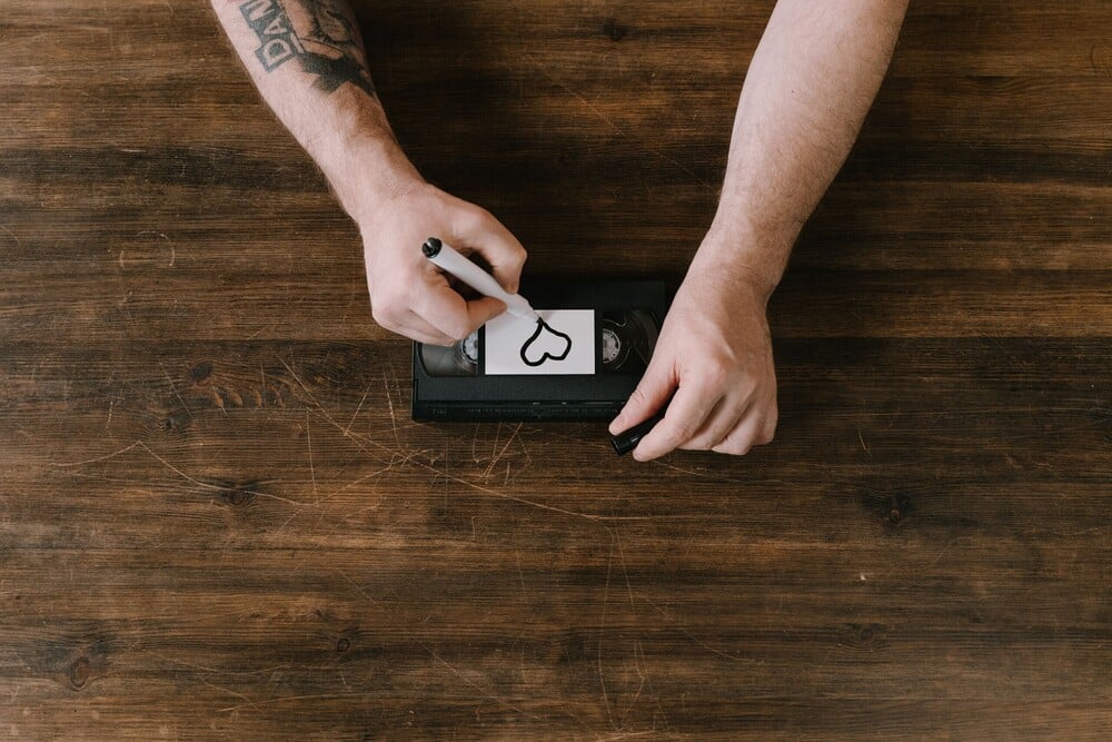 A person with tattoos writes a heart on a white label stuck to a VHS tape while sitting at a wooden table.