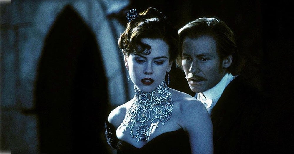 A woman in an ornate necklace and black dress stands in front of a man in formal attire. The scene is dimly lit, creating a dramatic and mysterious atmosphere.