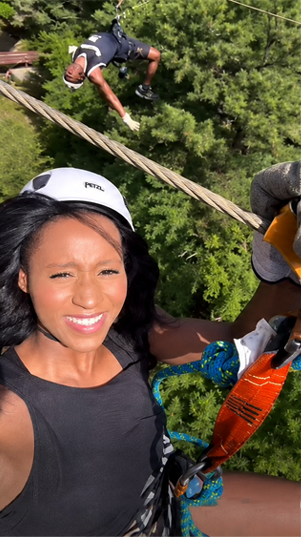 A woman wearing a helmet and harness smiles while zip lining above green trees. Another person is seen zip lining in the background.