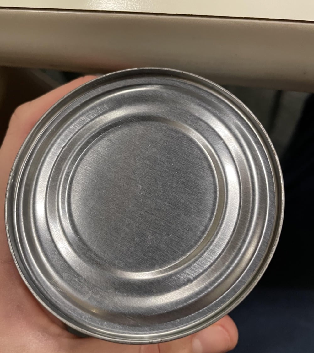 A hand holding a sealed, silver metal can with concentric ridges, viewed from above. The can is positioned near the edge of a light-colored table.