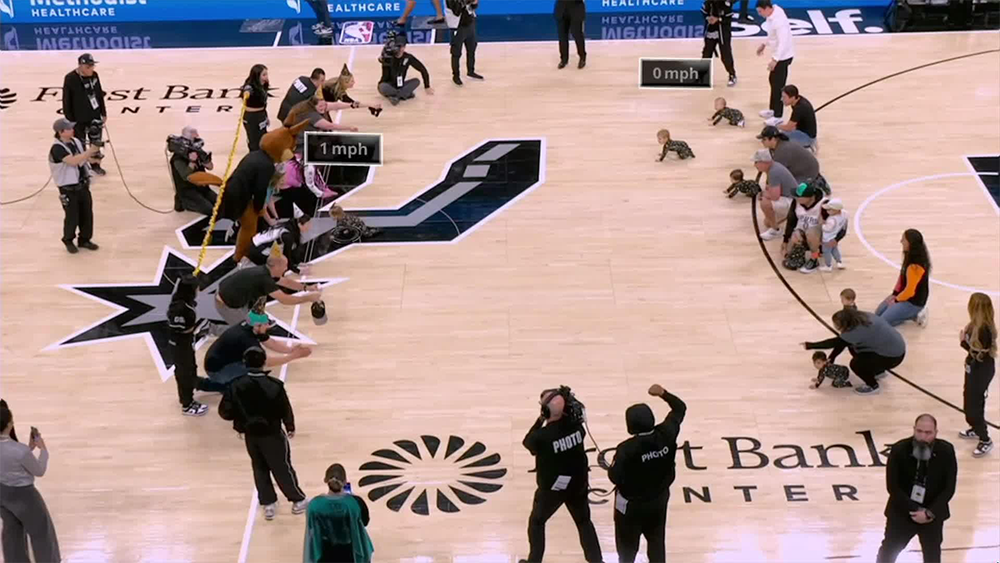Babies crawling in a race on a basketball court, surrounded by adults cheering and taking photos. Some speed measurements are shown on the screen, and photographers and officials are present.