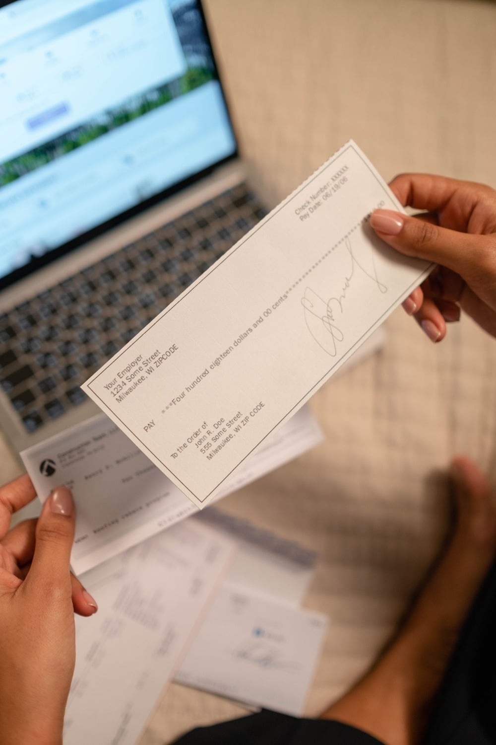 A person holding a paycheck above a laptop and some papers, with the check addressed to the individual. The background is slightly blurred, focusing on the hands and paycheck.