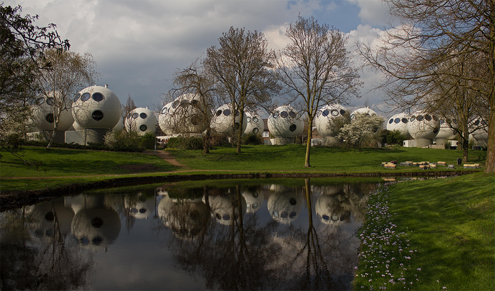 A row of white, spherical houses with round windows stands among leafless trees, reflected in a calm pond under a cloudy sky. Grass and flowers surround the scene.