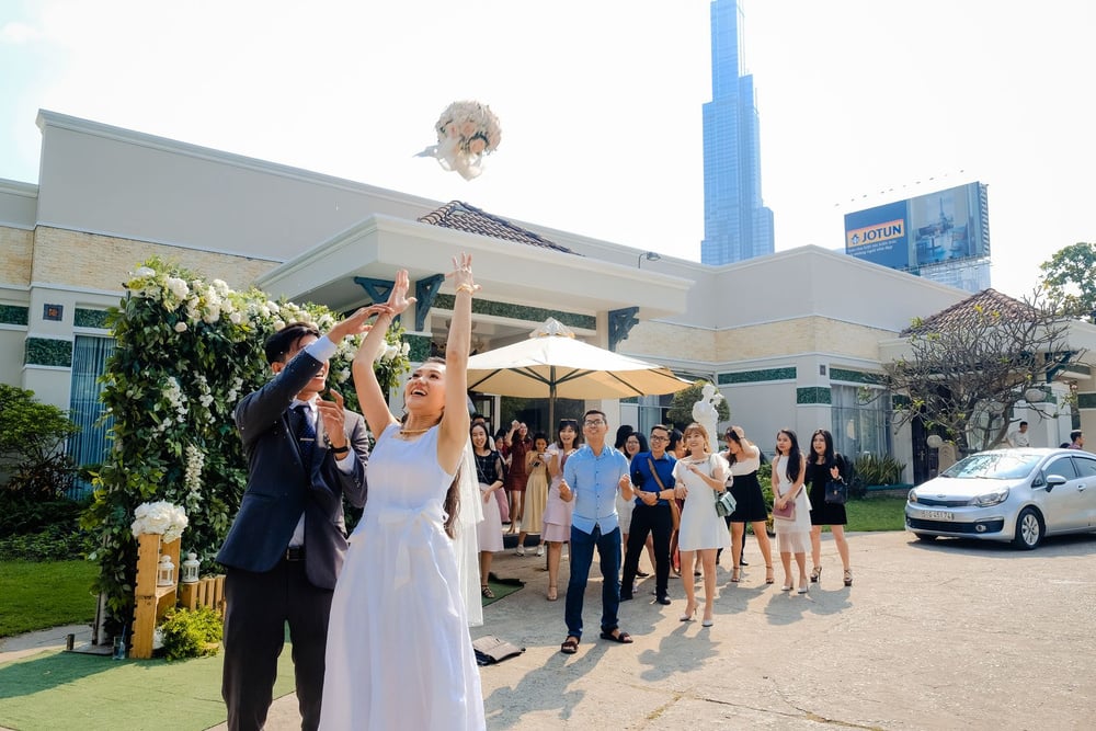 A bride tosses her bouquet while guests eagerly watch and reach to catch it, outside a wedding venue on a sunny day, with a tall skyscraper in the background.