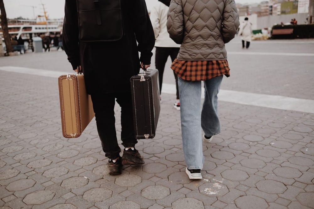 Two people walk on a paved street, each carrying a vintage suitcase. Both wear jackets and casual clothes. The image is taken from behind, and more people are visible in the background.
