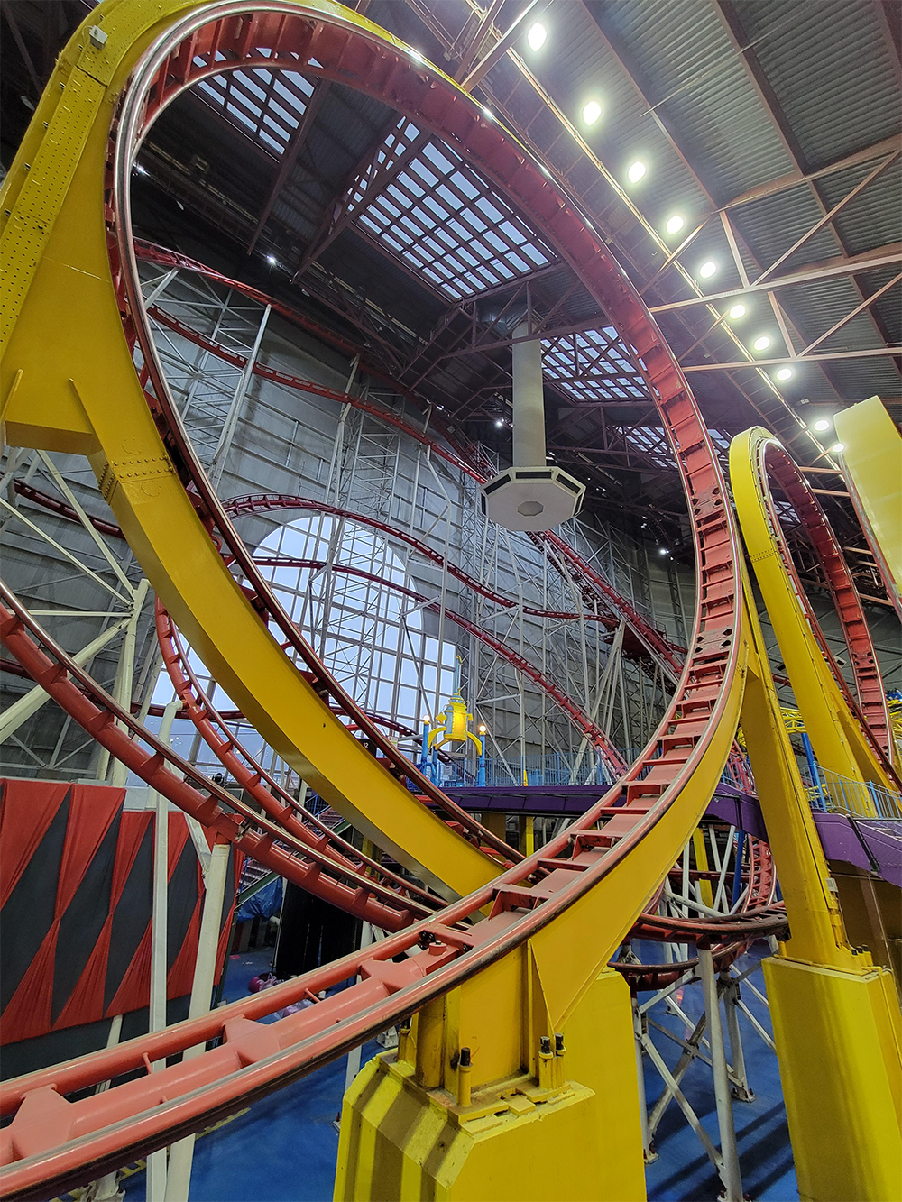 A yellow and red indoor roller coaster features a vertical loop, surrounded by metal beams and a high ceiling with lights. Blue flooring and structural elements are visible below the track.