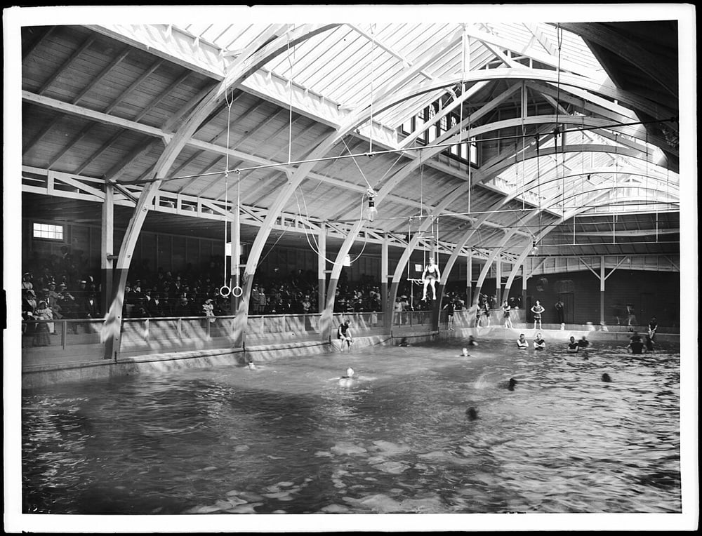 A large indoor swimming pool with high arched beams, a crowd watching from bleachers, and several people swimming or using gymnastic rings above the water.