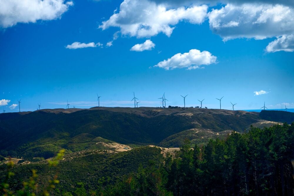 Wind turbines line the ridge of a green, hilly landscape under a bright blue sky with scattered clouds. Forested areas are visible in the foreground.