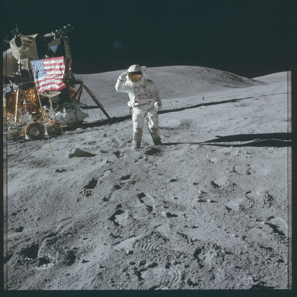 An astronaut in a spacesuit stands on the moon’s surface, saluting near an American flag and lunar module, with craters and hills visible in the background under a dark sky.