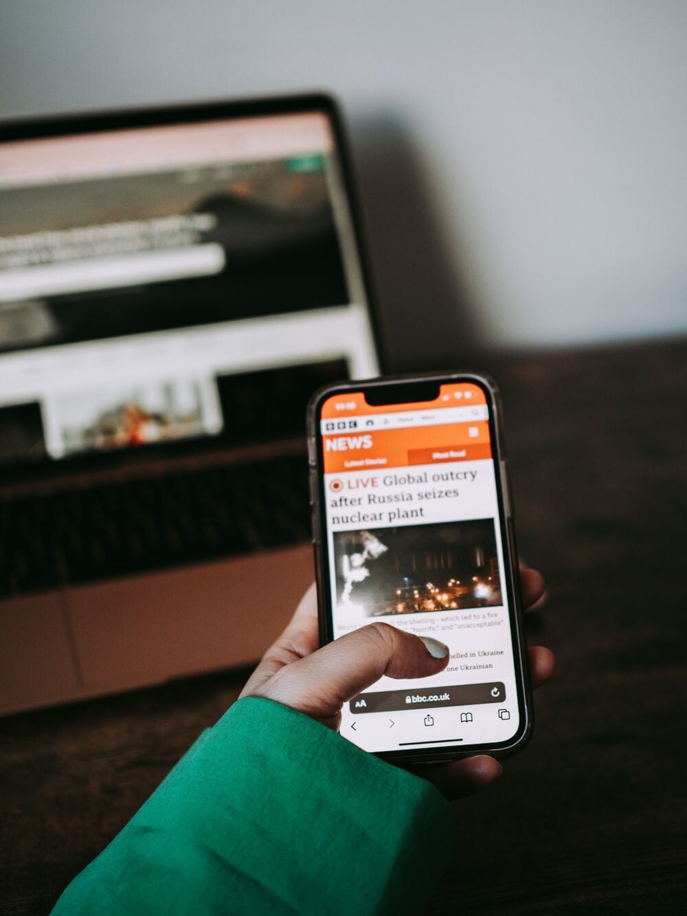 A person wearing a green sleeve holds a smartphone showing a news article about Russia seizing a nuclear plant. A laptop with a blurred website is in the background on a wooden table.