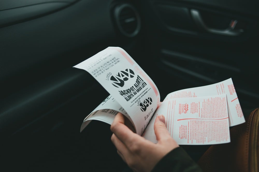 A person holding multiple lottery tickets with bold text and red print, sitting inside a car. The car’s door and dashboard are visible in the background.