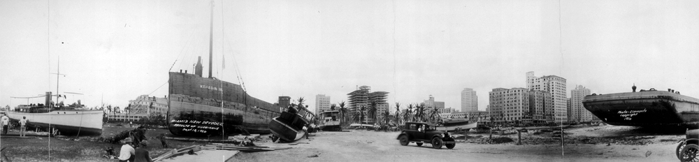 Black and white photo of ships and boats stranded onshore, cars and people nearby, with tall buildings and palm trees in the background; the scene appears chaotic, likely after a hurricane or natural disaster.