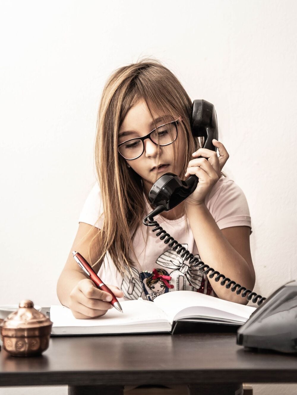 A young girl with glasses writes in a notebook while talking on a black corded phone, sitting at a desk with a copper container and a phone base.