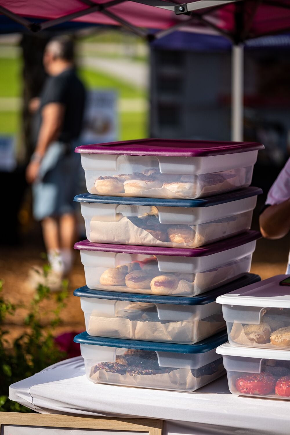 Four stacked plastic containers filled with bagels and pastries are on an outdoor table, with a blurred figure and greenery in the background.