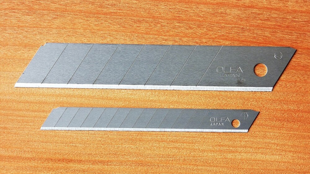 Two silver utility knife blades of different sizes lie parallel on a wooden surface. Each blade has segmented snap-off edges and a hole at one end. The brand “OLFA JAPAN” is visible on both blades.