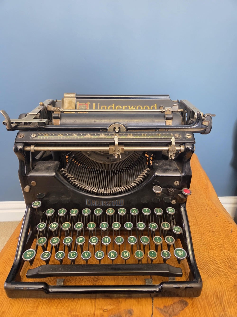 A vintage black Underwood typewriter sits on a wooden desk against a blue wall, with round keys and visible metal components.