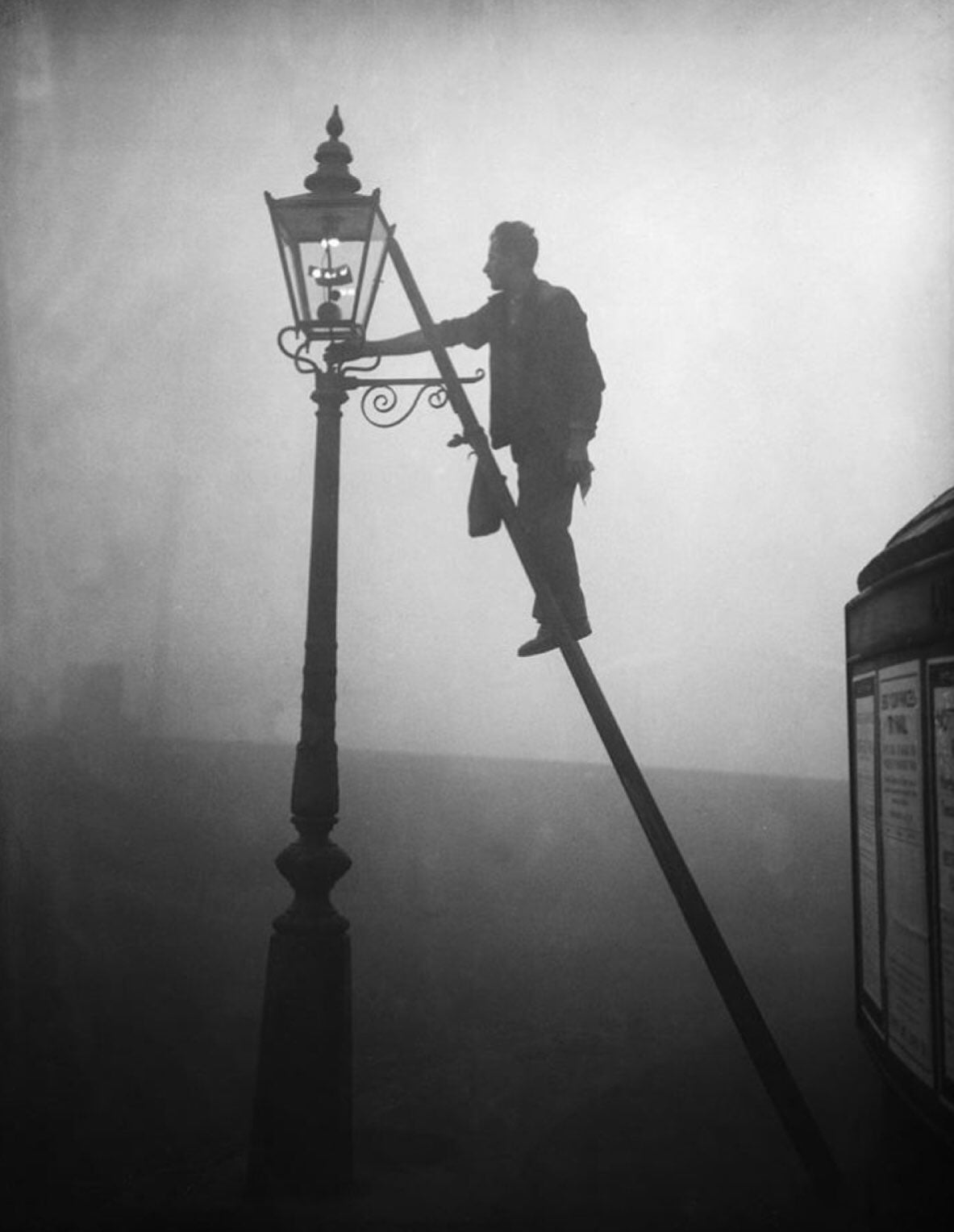 A person stands on a ladder, lighting or maintaining a gas streetlamp in thick fog. The scene is atmospheric, with silhouettes and low visibility creating a moody, vintage feel.