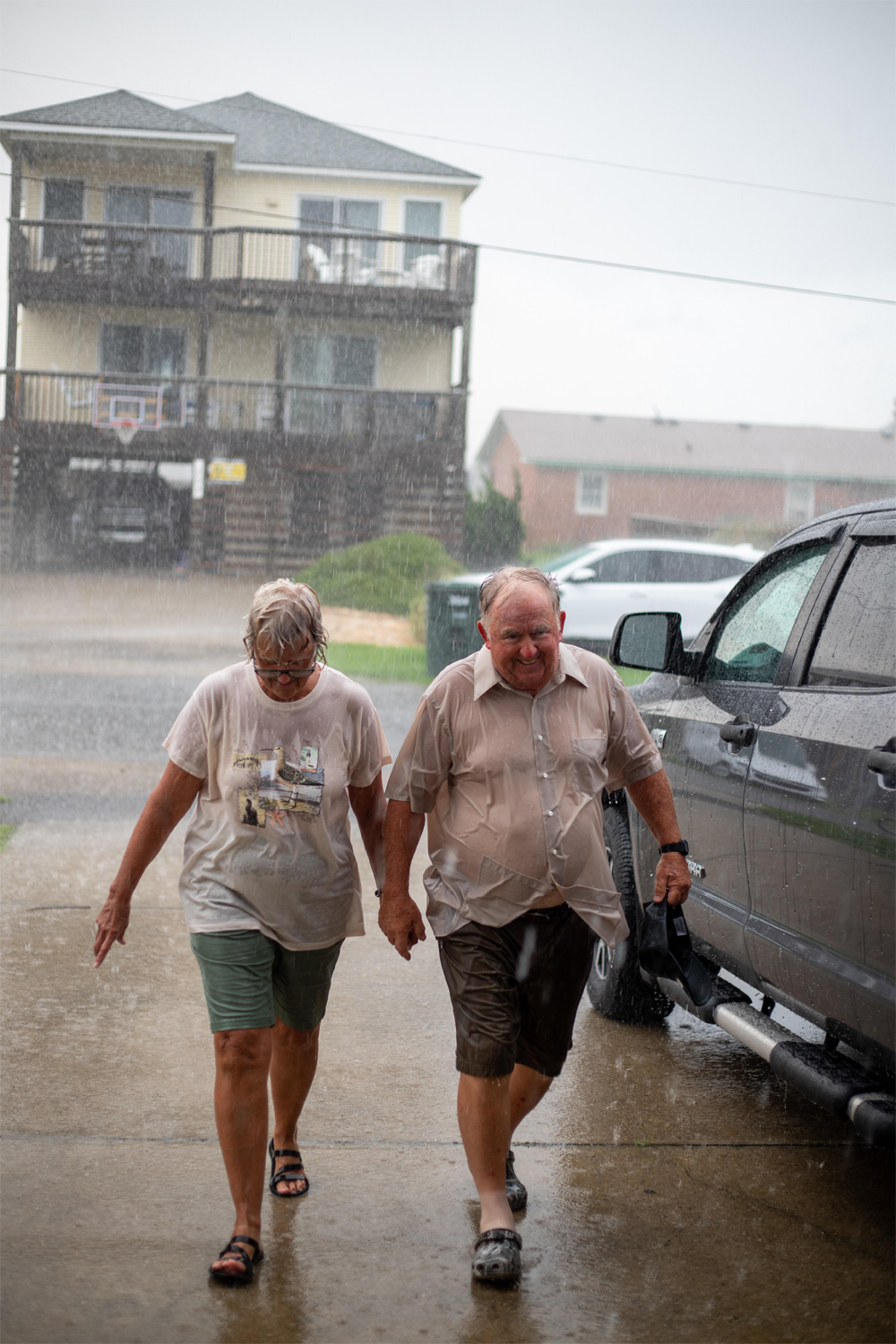 An older couple holding hands walks in the rain toward a car, looking down and wet. Houses and parked vehicles are visible in the background.