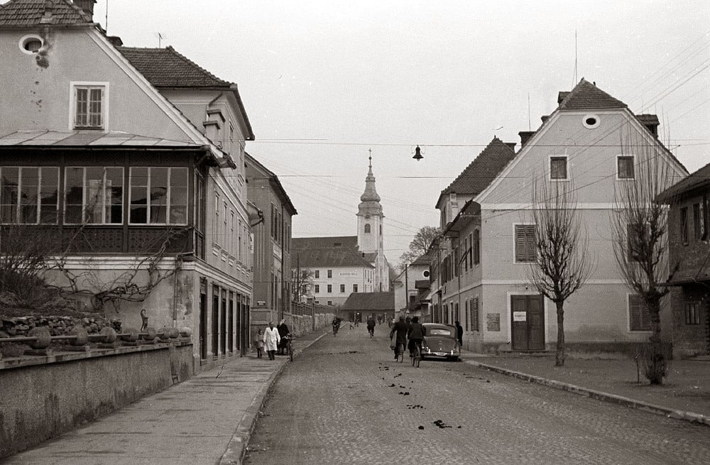 Black-and-white photo of a cobblestone street lined with old buildings, a church with a tall steeple in the background, people walking, and a classic car parked along the road.