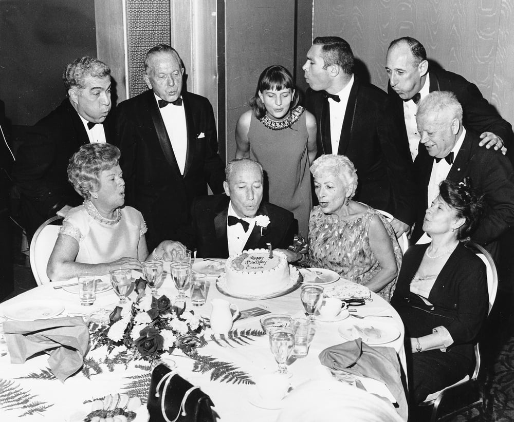 A group of men and women in formal attire gather around a table, watching an elderly man blow out candles on a birthday cake. The mood appears celebratory, and the setting is an elegant banquet or party.