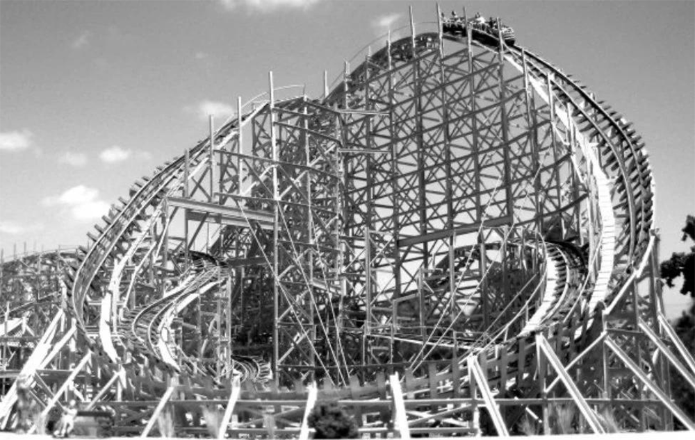 A black-and-white photo of a wooden roller coaster with looping tracks and riders at the top, surrounded by a support structure against a partly cloudy sky.