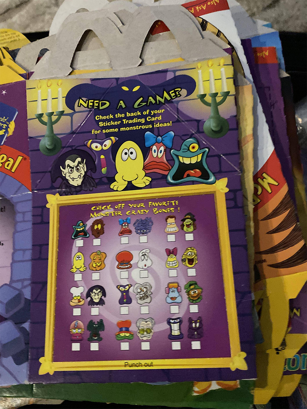Colorful McDonald’s Happy Meal box featuring cartoon monster characters, a punch-out game with 20 monster faces, and instructions to check off favorites. Background includes other boxes stacked behind.
