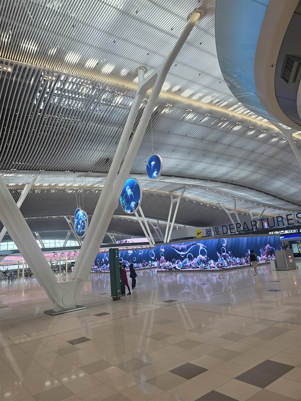 Modern airport terminal with a high, curved ceiling, digital globe displays, large “DEPARTURES” sign, and a digital aquarium wall. Few people and luggage are visible on the shiny tiled floor.