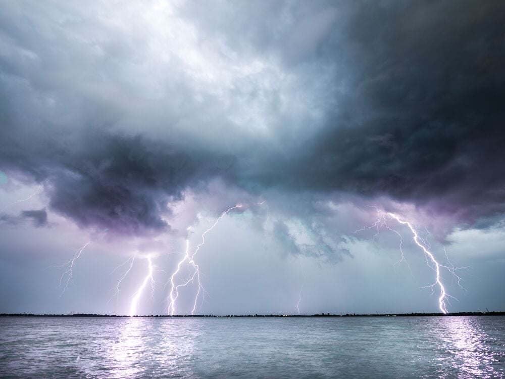 Lightning bolts strike over a body of water under dark, stormy clouds. The sky is illuminated by the flashes, and the water reflects some of the light. The scene appears intense and dramatic.