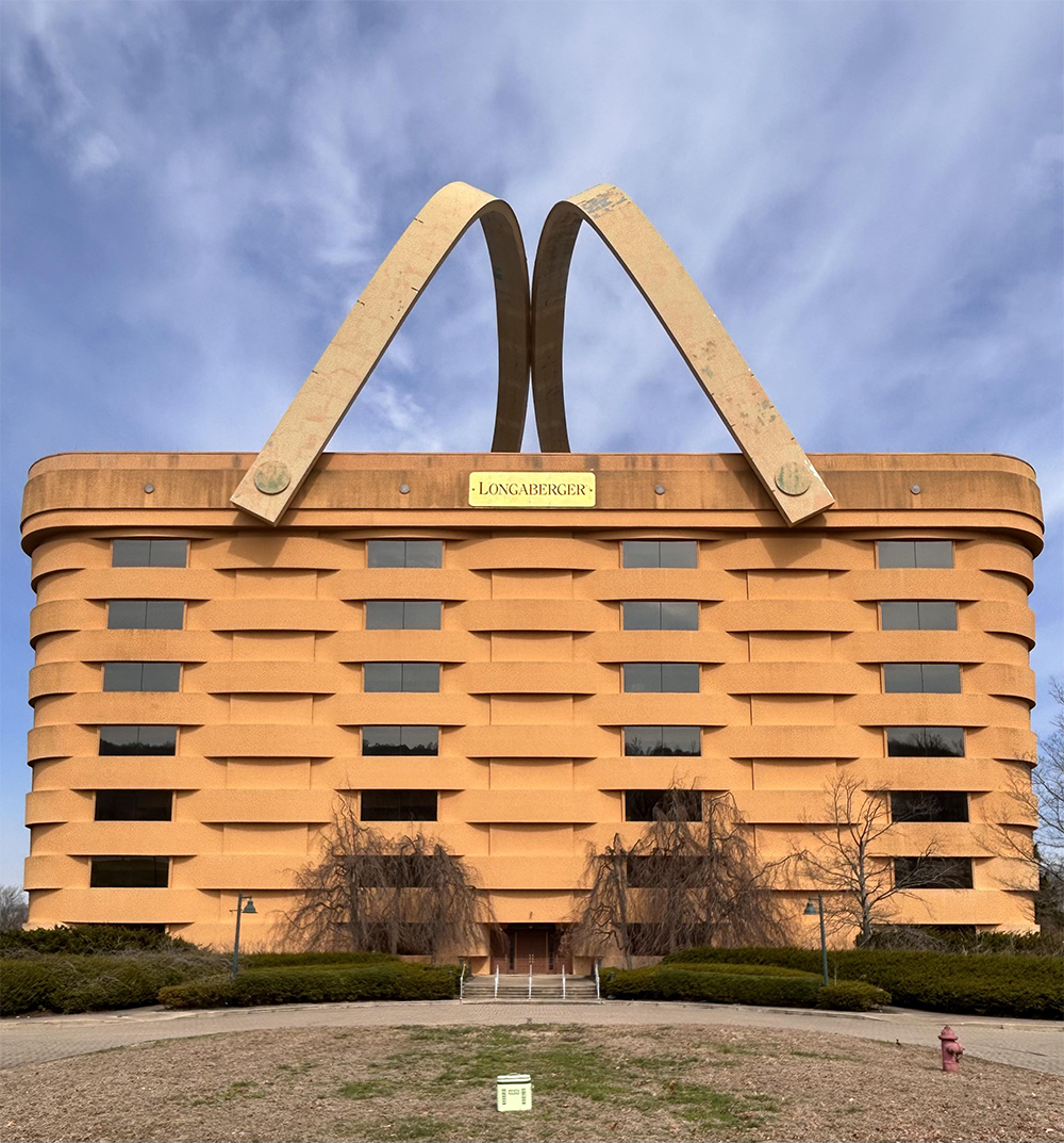 A seven-story office building shaped like a giant woven basket with two large handles on top, set against a blue sky. The sign reads “Longaberger.” Trees and bushes are visible in front.