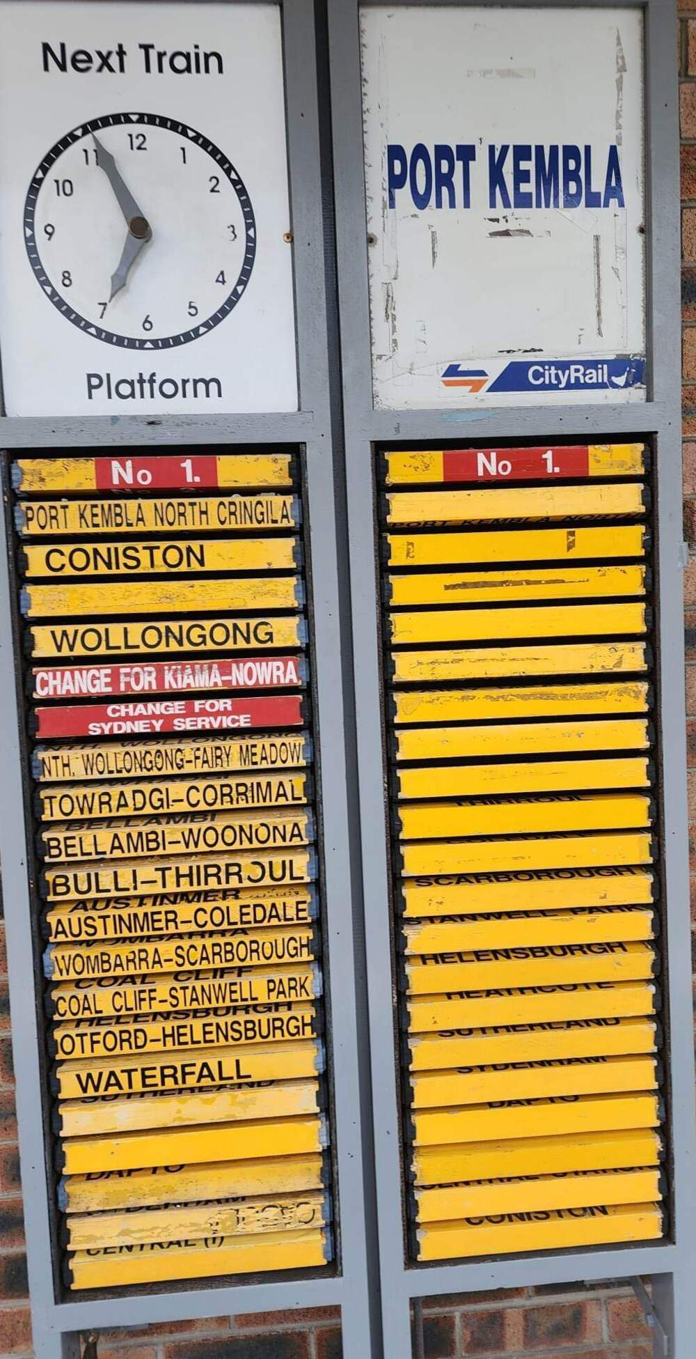 Train station sign showing the next train to Port Kembla from platform 1. Below are yellow signs listing stations including Coniston, Wollongong, Towradgi, Woonona, Thirroul, and Waterfall.