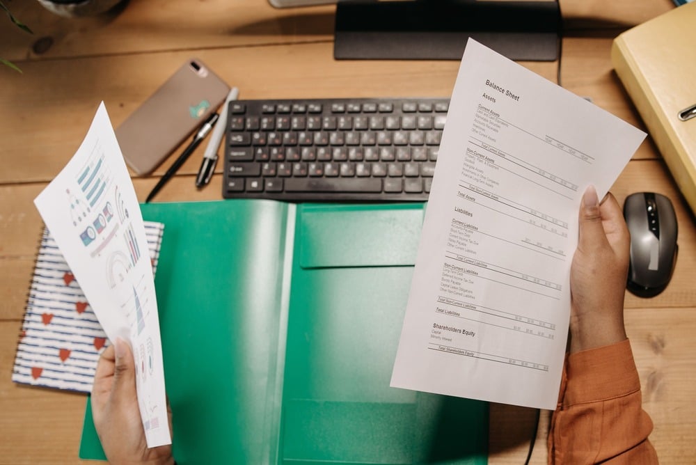 A person holding financial documents at a desk with a keyboard, phone, pen, computer mouse, and a green folder.