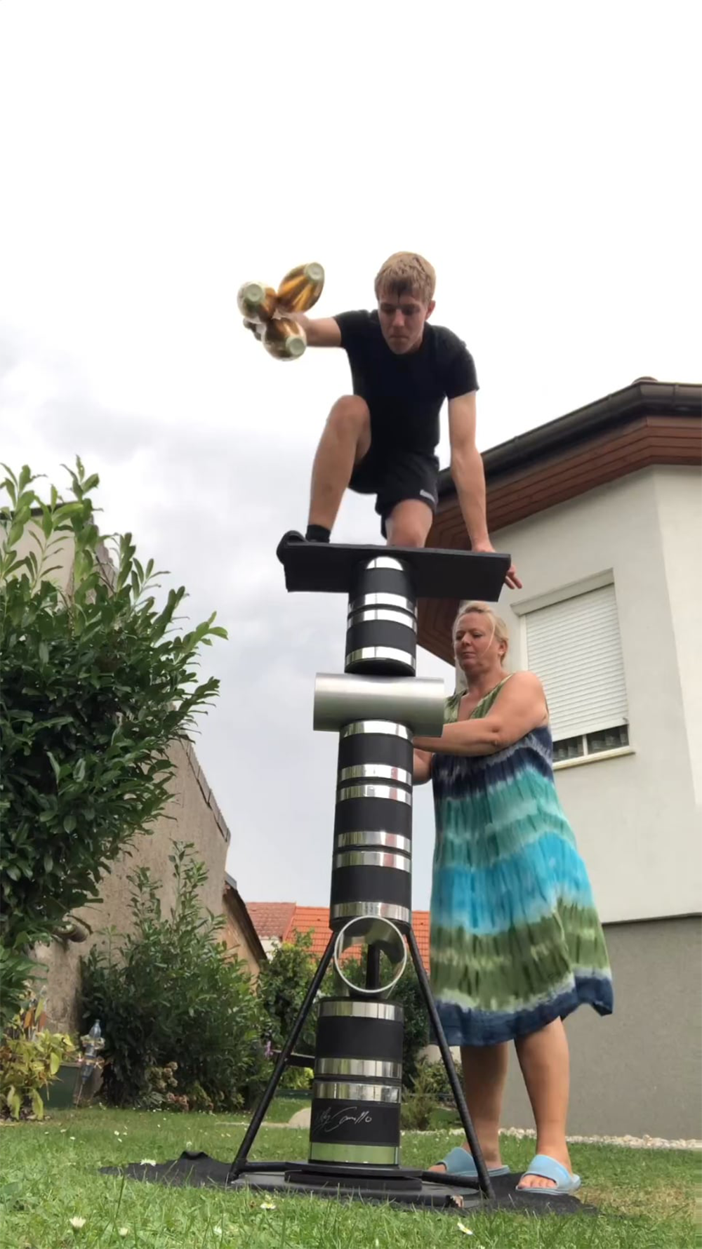 A woman steadies a tall stack of objects while a man kneels on a board at the top, holding bread. They are outside near two houses on grass, under a cloudy sky.