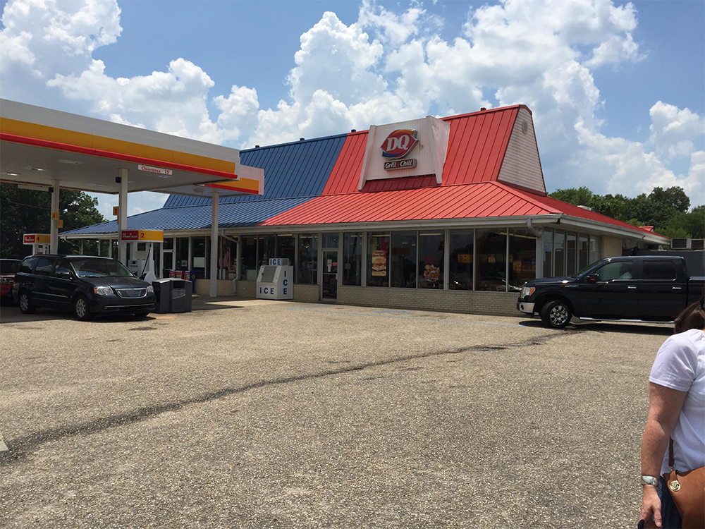 A Dairy Queen restaurant with a red and blue roof is next to a Shell gas station. Several cars are parked outside, and bags of ice are stacked near the entrance. A partly cloudy sky is visible above.