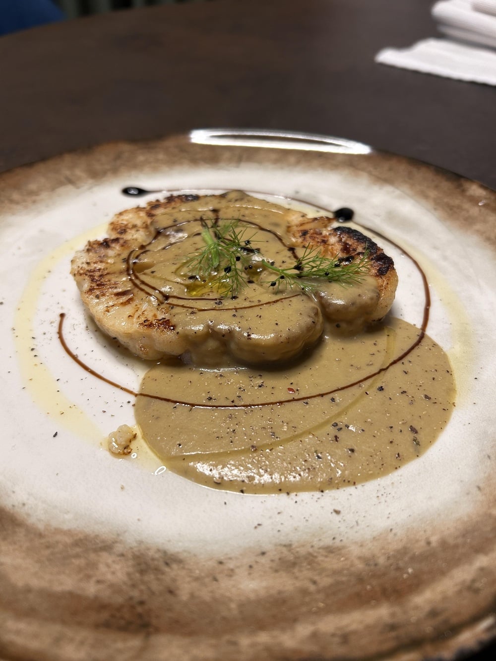 A grilled cauliflower steak topped with creamy sauce, garnished with fresh herbs, sits on a rustic plate with artistic balsamic and oil drizzles.