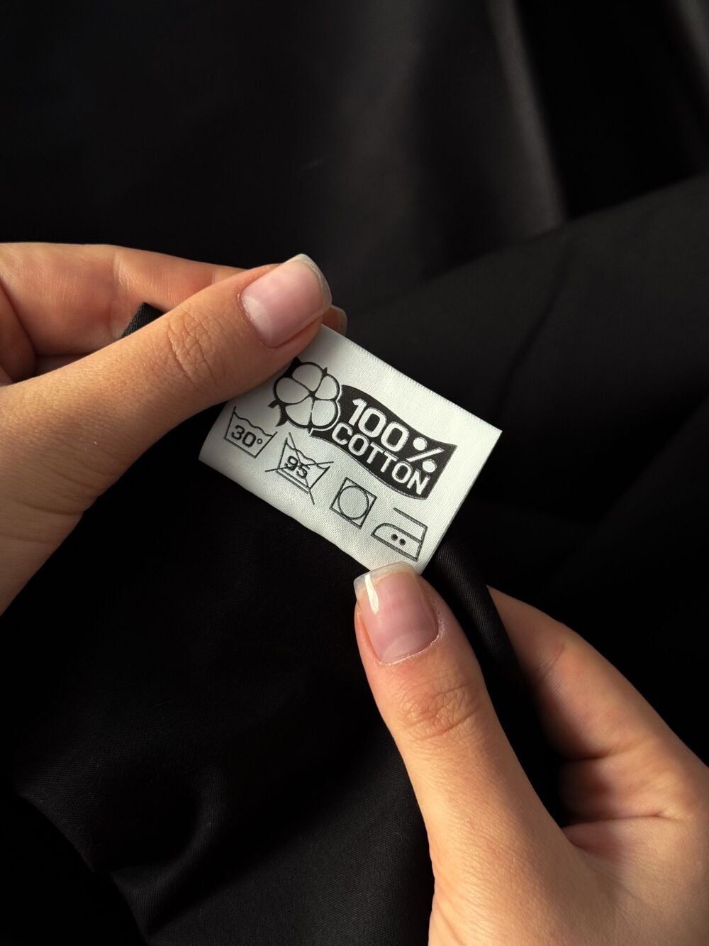 Two hands hold a clothing label on black fabric. The label reads “100% cotton” and displays laundry symbols for washing at 30°C, no bleach, tumble dry, and iron at medium heat.