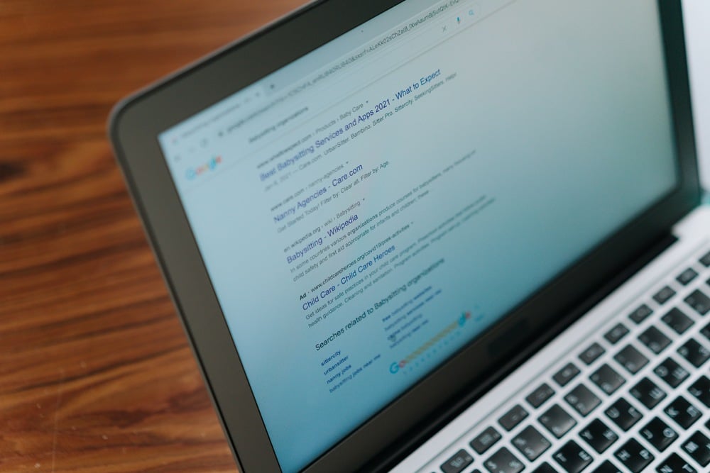 A laptop on a wooden table displays a Google search results page about best babysitting services and related topics.
