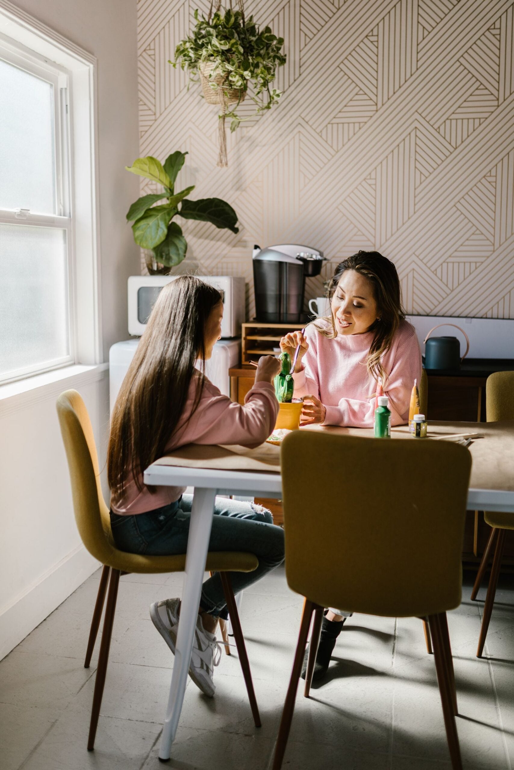 Two people sit at a kitchen table, smiling and painting small green bottles. Sunlight streams through a window, and plants decorate the modern, patterned room.
