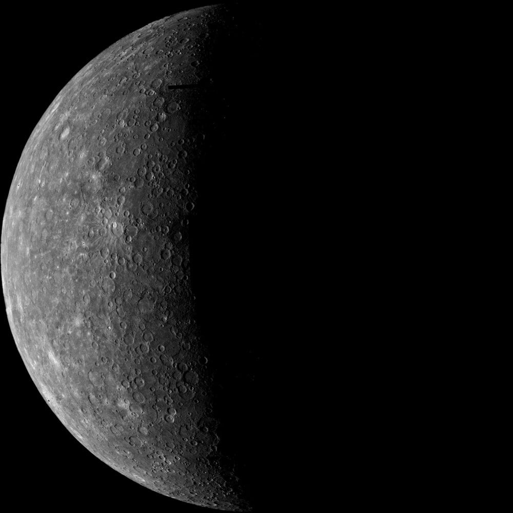 A grayscale image of a cratered, rocky planet half illuminated, with the other half in shadow, against a black background. The surface is covered in circular craters of varying sizes.