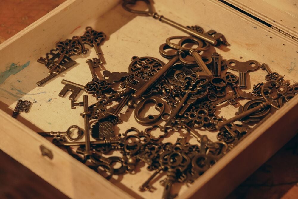 A wooden box filled with various ornate, vintage-style metal keys and keyholes, scattered inside on a light-colored surface.