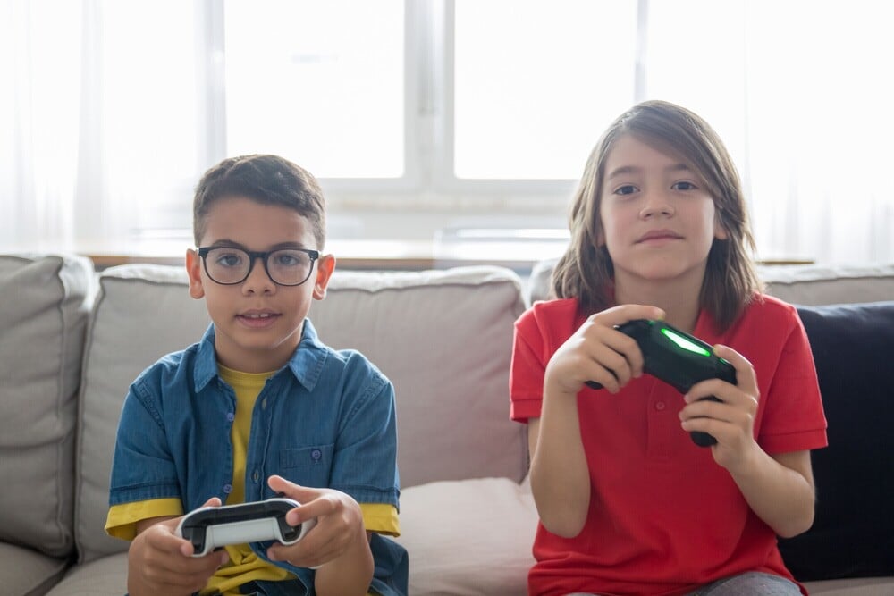 Two kids sit on a couch playing video games, each holding a controller. One wears glasses and a blue shirt, the other has long hair and a red shirt. They appear focused, with a bright window in the background.