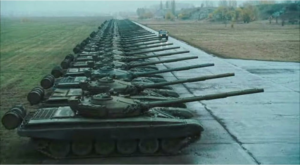 A long row of military tanks is lined up on a paved surface beside a grassy field, with a small vehicle parked near the tanks in the distance under an overcast sky.
