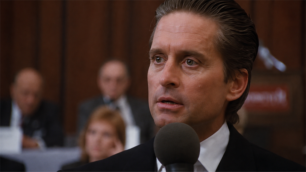 A man in a suit speaks into a microphone at a formal event, with blurred attendees seated in the background.