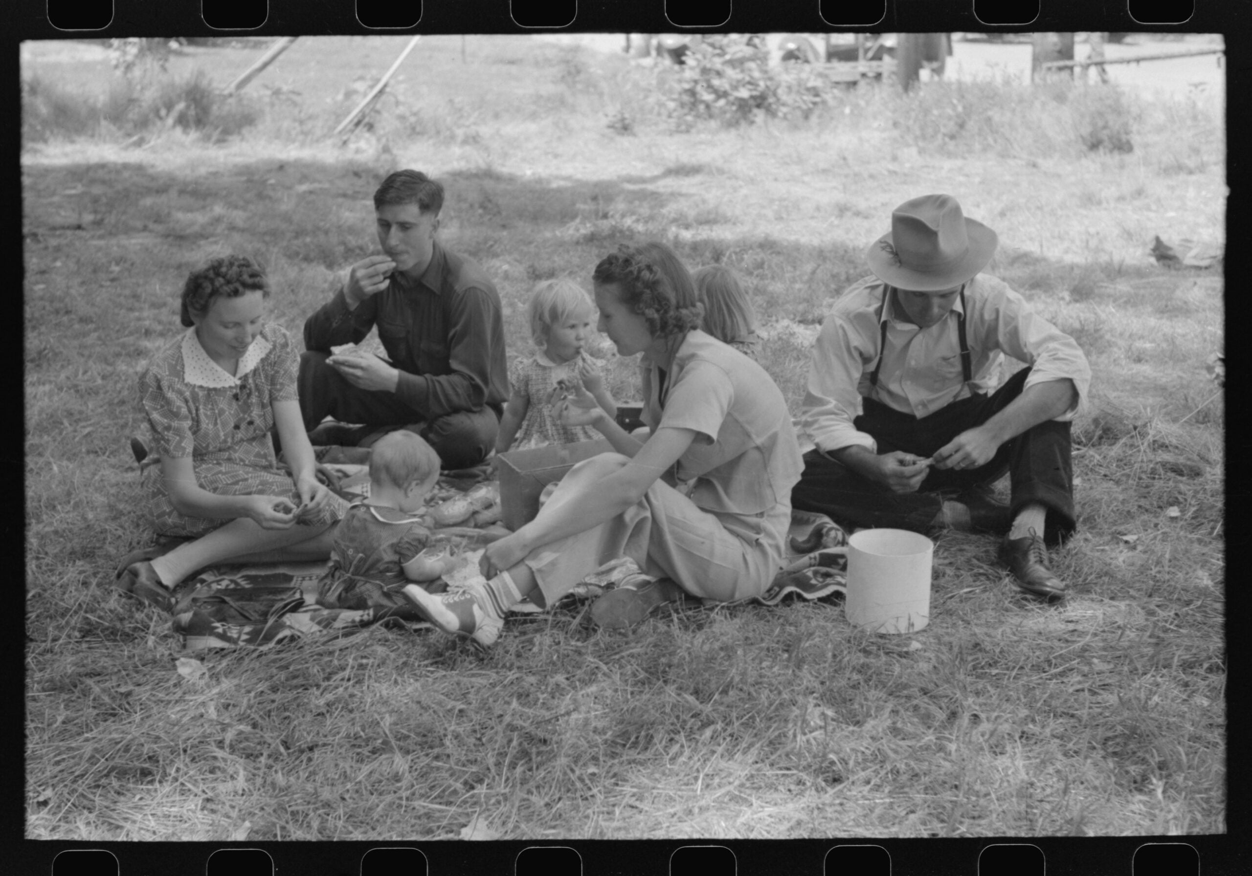 A group of adults and children sit on blankets in the shade having a picnic, sharing food and spending time together on a grassy area outdoors.