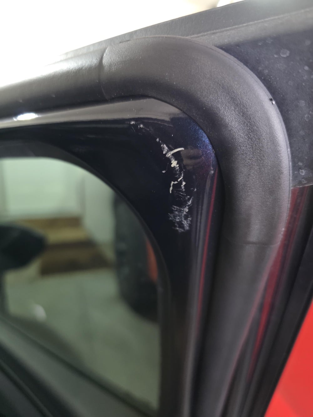 Close-up of a car door showing a small scratch with white marks on the black trim near the window. The background is blurry, with a garage door and floor faintly visible.